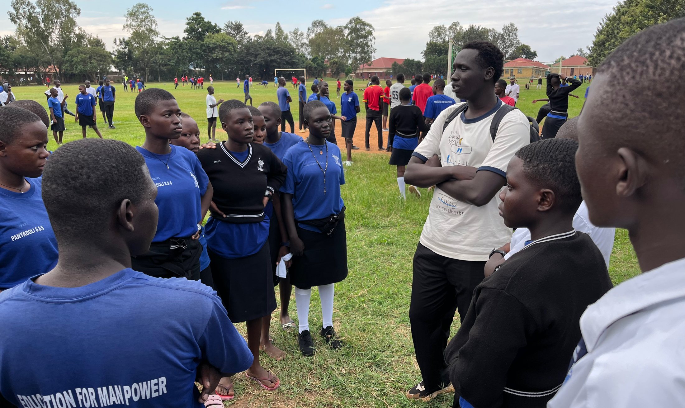 A young leader stands on a school field talking with a group of students in uniform who listen closely