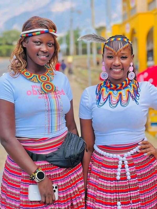 Two smiling young women stand side by side in beaded necklaces, headbands and bright striped skirts