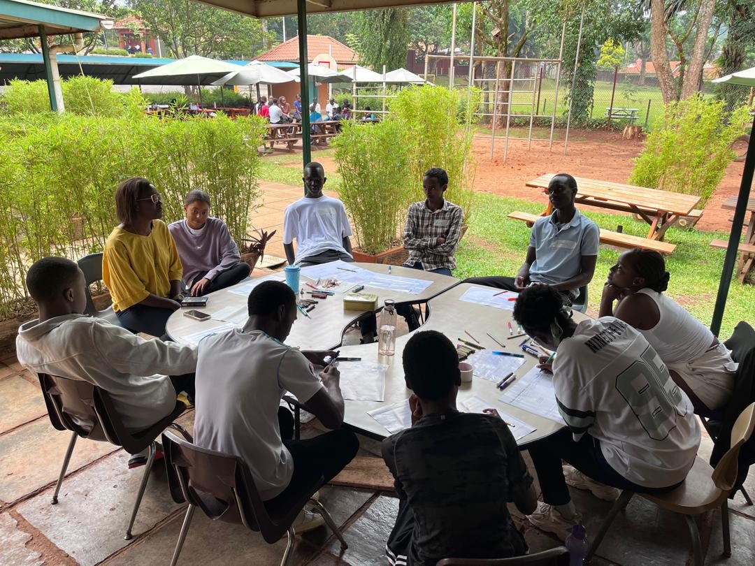 Young people seated around a table outdoors in discussion, with pens and paper in front of them