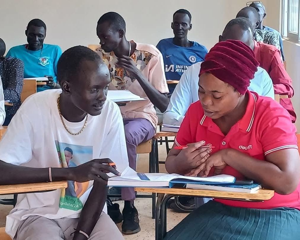 Two young adults work through a book together at a desk, with other students studying behind them