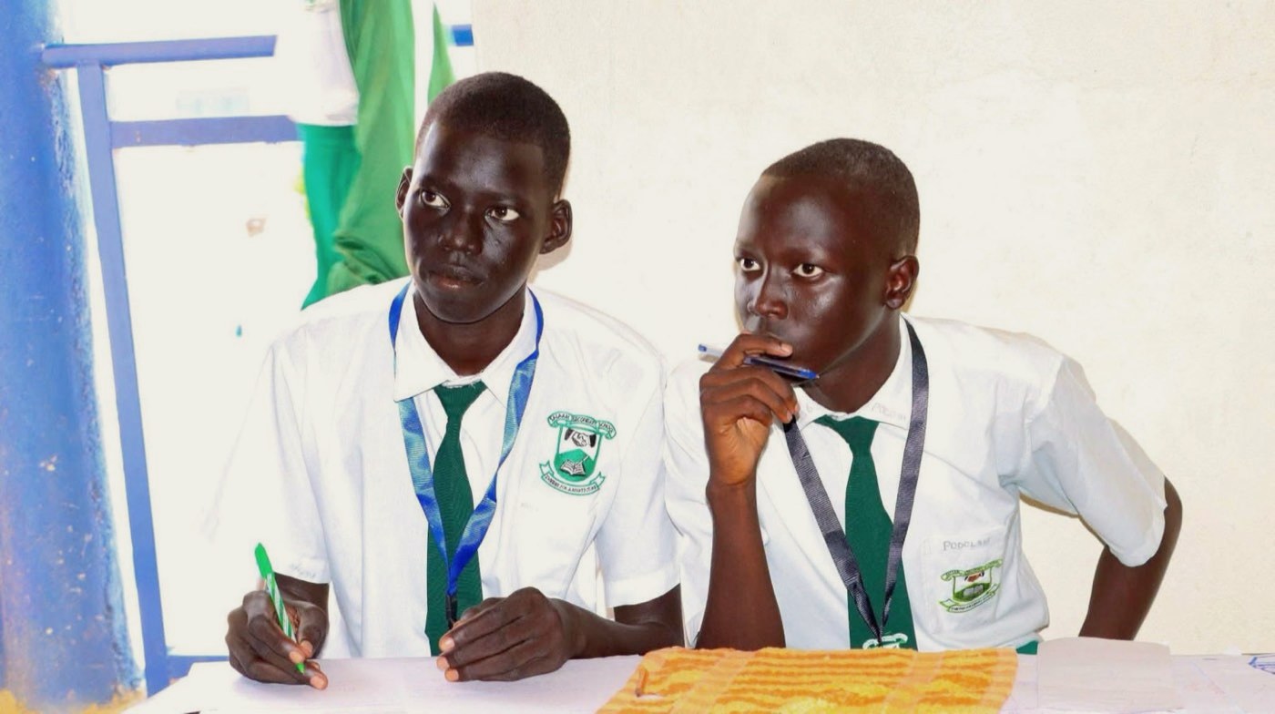 Two secondary school students in white shirts and green ties follow a classroom session, one taking notes and one listening with a pen in hand