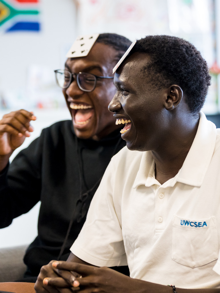 Two young men laugh together during a classroom game, one wearing a UWC South East Asia polo shirt